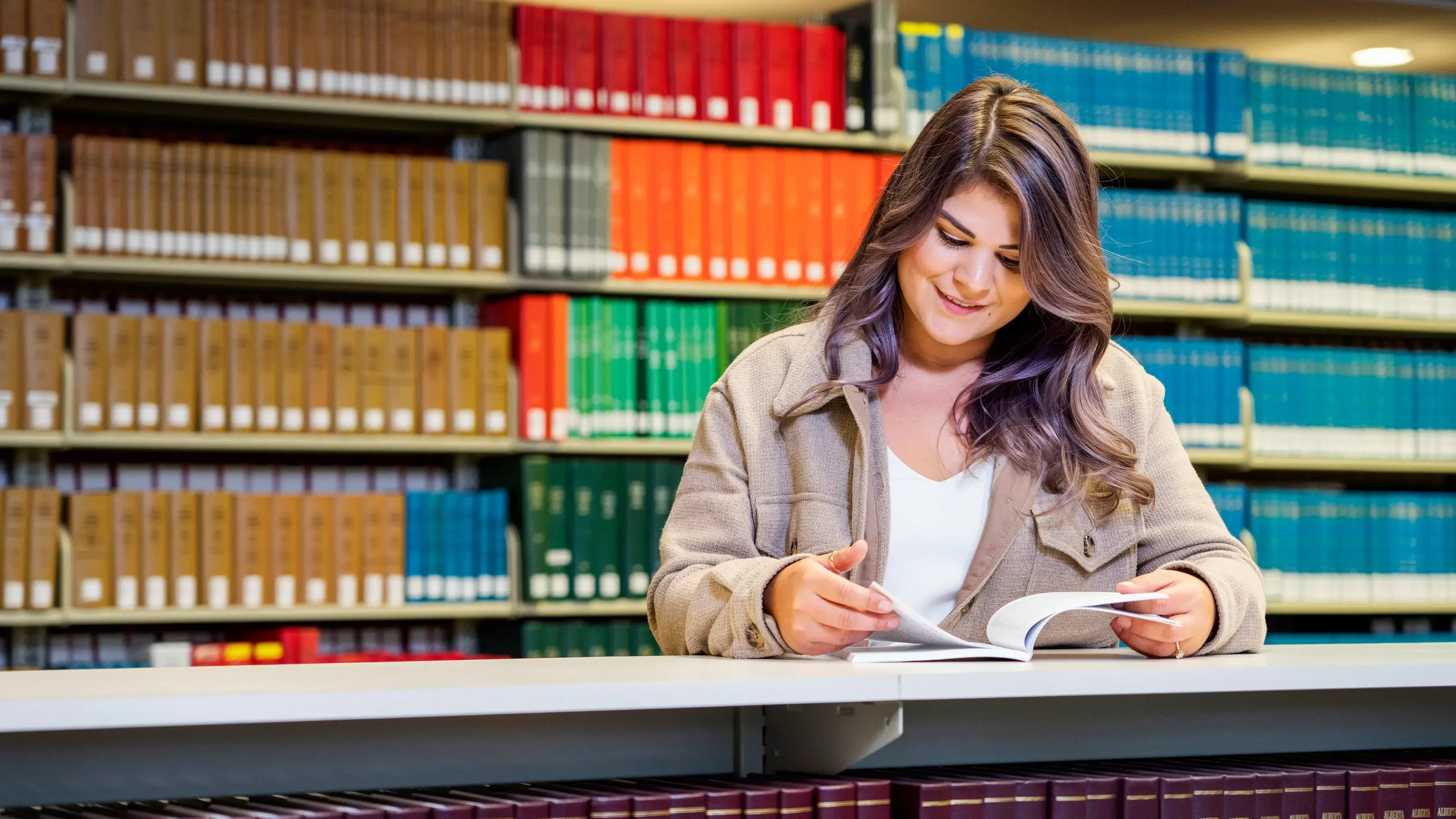 A woman smiles as she looks at a book with a bookshelf behind her.