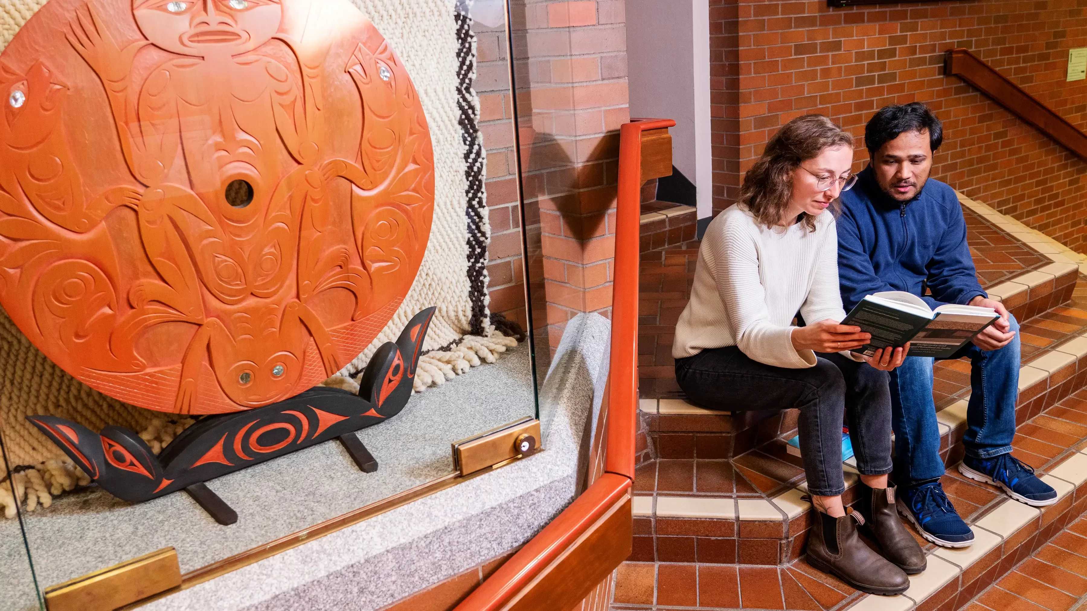Two students sit on stairs reading a book with indigenous art in foreground.