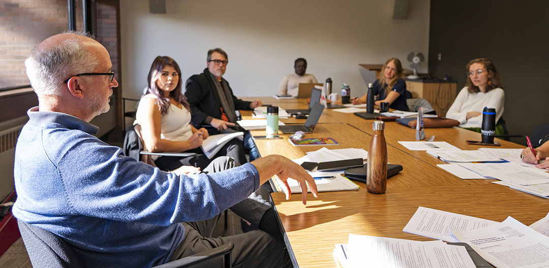 A group of people sit in a room at a large table scattered with papers