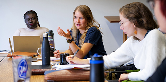 Three sudents sit at a long table with laptops and papers