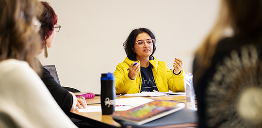 A student in a yellow shirt talks to other students around a table