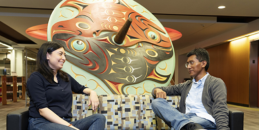 Two graduate students talk while sitting on a couch in front of a large, carved spindle whorl.
