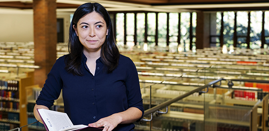 A graduate student stands on the mezzanine of the library holding a book