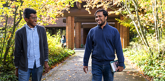 Two students walk on a sidewalk in the trees, laughing.