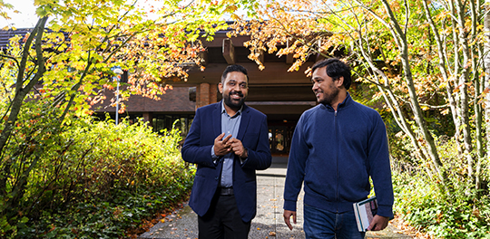 Two students walk along a sidewalk in the trees, talking.