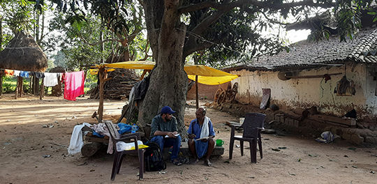 A student sits next to a man outside of his dwelling in India