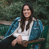 A photo of Asia Youngman. Asia is sitting on a bench outdoors. She is smiling and her hands are crossed. A denim jacket is draper over her shoulders. She has long brown hair that falls over her shoulders. 