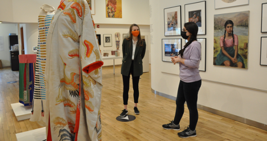 Two students wearing face masks stand in Legacy Art Gallery Downtown, discussing the artwork on display.