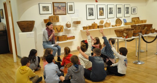 a group of students gazes at one of Legacy's educators. Some of the students have their hands raised and are ready with answers to the question they were asked. In the background there are baskets mounted on the gallery's walls. 