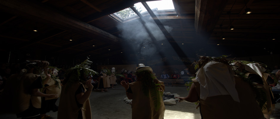 Two figures in wood masks, wrapped in greenery, mid-dance. 