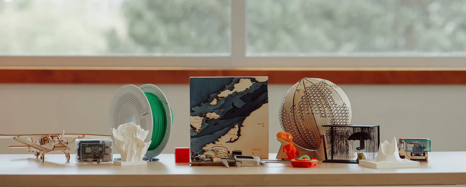 Laser cut and 3D printed objects arranged in front of a large window with trees in the background.