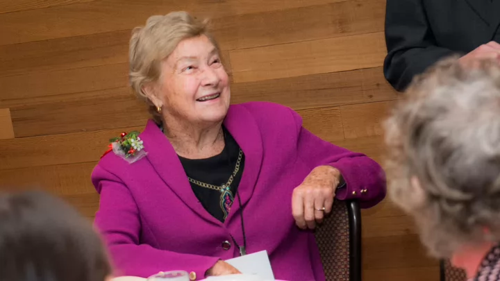 Joyce is wearung a fuscia jacket with a flower corsage on one shoulder.