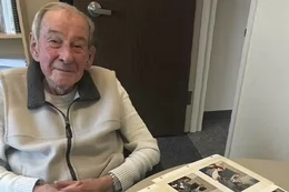 Werner is wearing a white sweater with a zipper sitting at a table with a photo album in front of him.