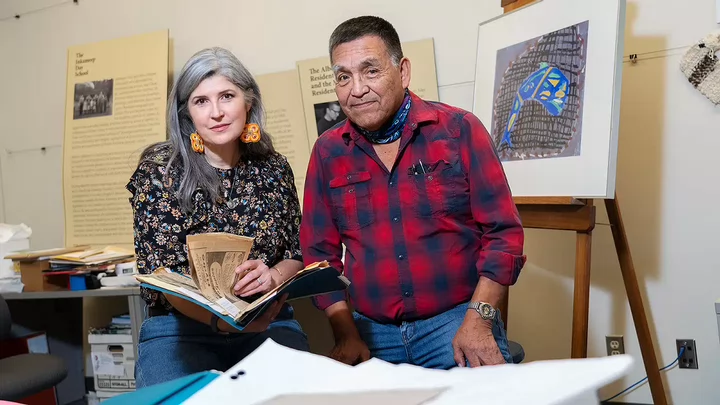 Andrea and Mark holding research documents in front of posters about residential schools and framed artwork.
