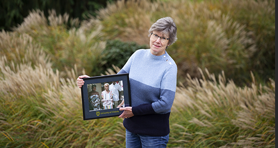 Ann standing amongst tall grass behind Mearns library holding a framed photo