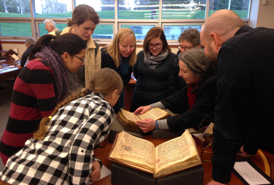 A group of about ten people arranged in a circle looking at two manuscripts with decorative text elements.