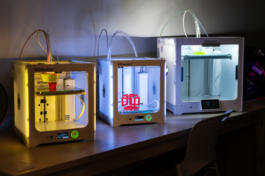 Three 3D printers sitting on a desk.