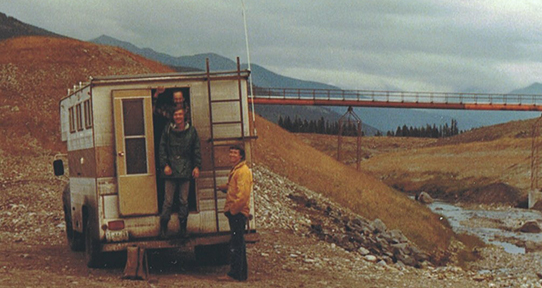 Two men standing near a white truck beside a river bed and a bridge in the background