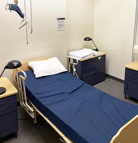Respite room with adjustable single bed with lift, spare sheets, desk lamps, and side tables with drawers.