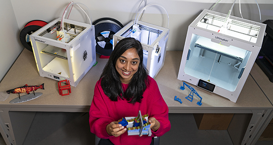 Sowmya wearing a red top sitting in front of three 3D printers