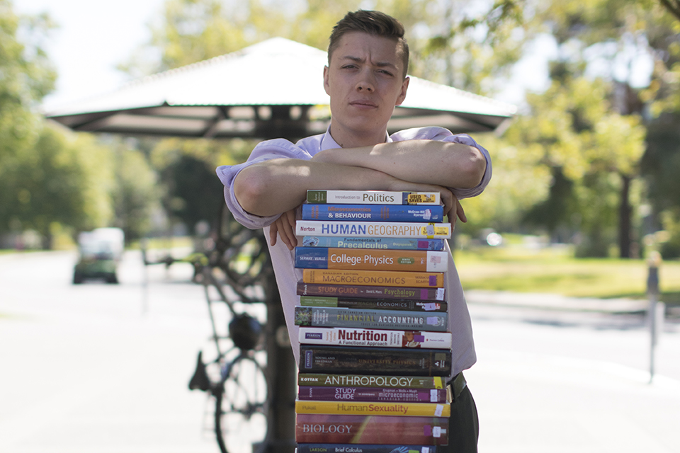 Maxwell Nicholson stands in front of a large stack of textbooks. His arms are crossed over the top of the stack.