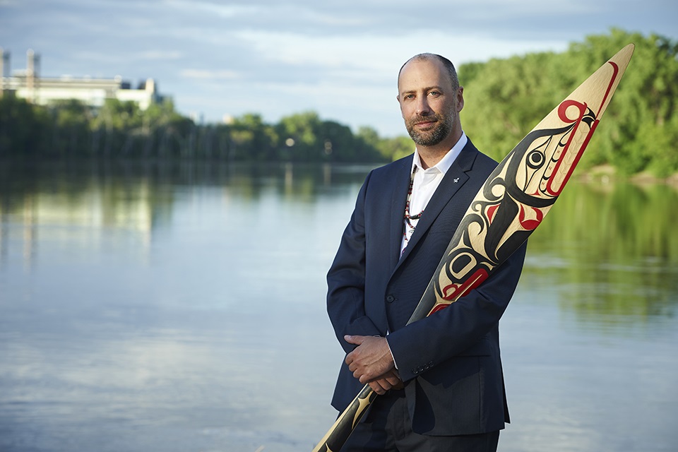 Ry Moran, along the banks of the Red River in Winnipeg, holding one of three reconciliation paddles carved by artist Carey Newman and designed by artists from different generations and nations for the 2019 Building Reconciliation forum. Credit: Nardella Photography