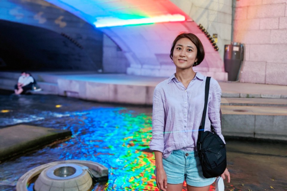 Sujin Lee pictured in a purple shirt and standing near water with a white brick wall behind her.