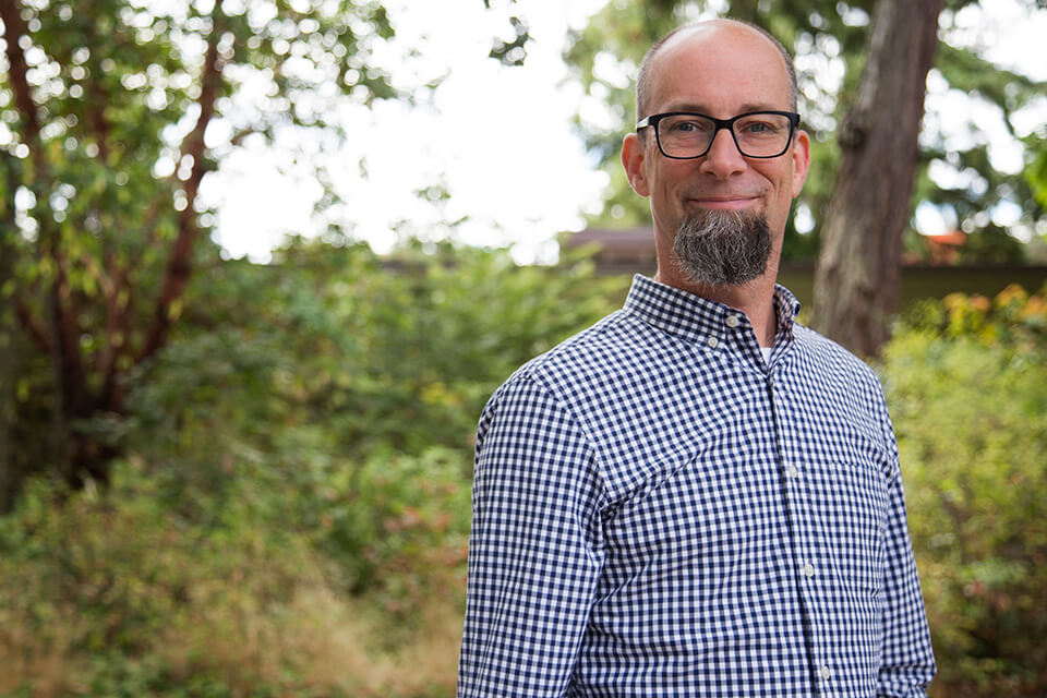 Jan Kietzmann stands in front of a green bushy forest. He is wearing a black and white plaid dress shirt and is smiling at the camera.