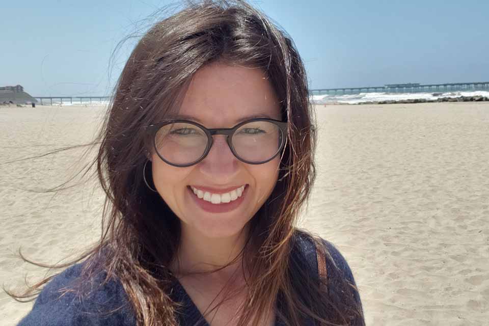 UVic PhD graduate smiles on a sandy beach in front of crashing waves on the shore.