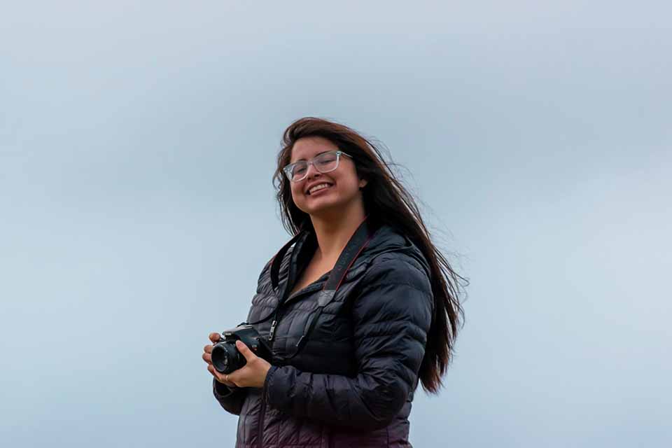 UVic writing grad Jenessa Joy Klukas stands in front of a grey-blue sky, holding a camera and smiling brightly.