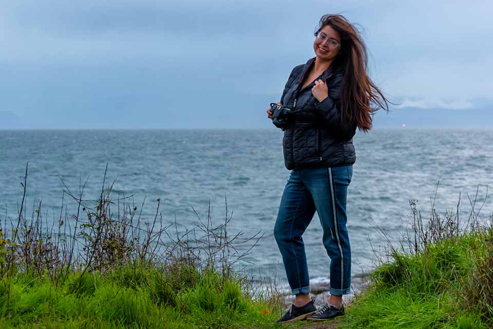 UVic writing grad Jenessa Joy Klukas poses with her camera on a grassy hill in front of the ocean.