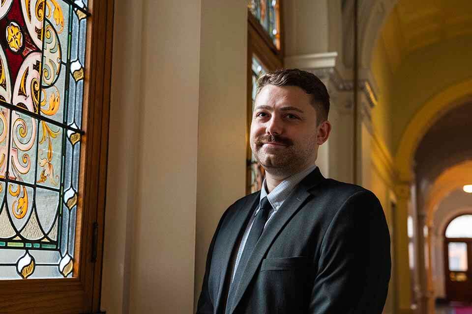 UVic Public Administration grad Matthew Creswick poses in a suit and tie in front of a stained glass window.