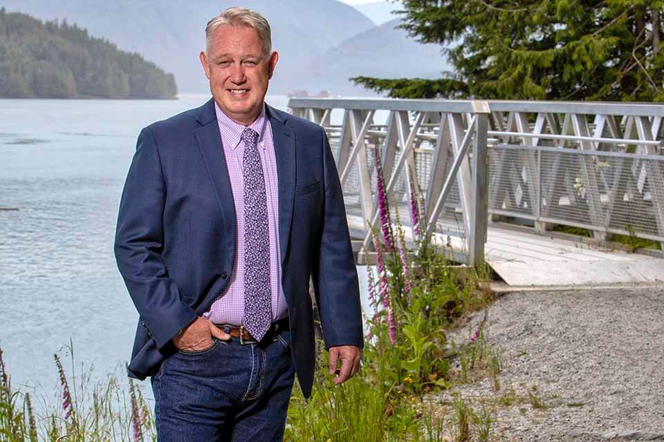 Cory Stephens, recipient of the Award for Excellence in Aboriginal Relations, stands smiling in front of a dock by the water. He wears a suit jacket, tie, and a pair of jeans.