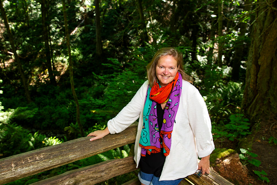 Heather Castleden stands in front of a green forested area, leaning against a log railing.