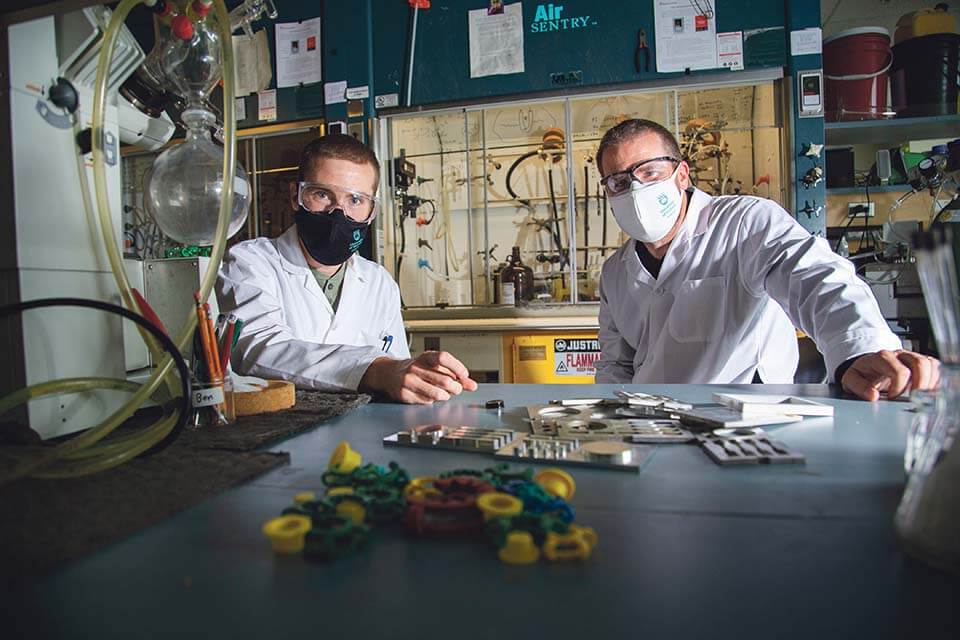 Ben Godwin and Jermey Wulff stand in their lab around a table with auto parts laid out on it. They wear protective goggles and UVic face masks.