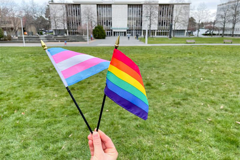 A hand holds up a Pride flag and a Trans Pride flag in front of the McPherson Library