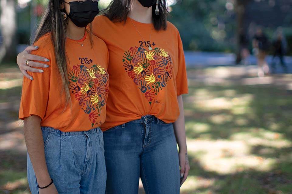 Two UVic students stand with their arms around each other. Each is wearing an Orange Shirt Day t-shirt with Carey Newman's