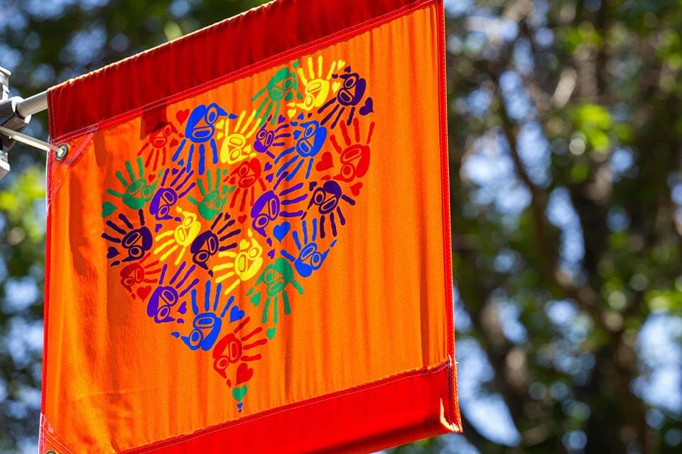 An orange flag with a heart made out of rainbow coloured children's' hand-prints hangs from a horizontal pole. The graphic was designed by Carey Newman for Orange Shirt Day. 