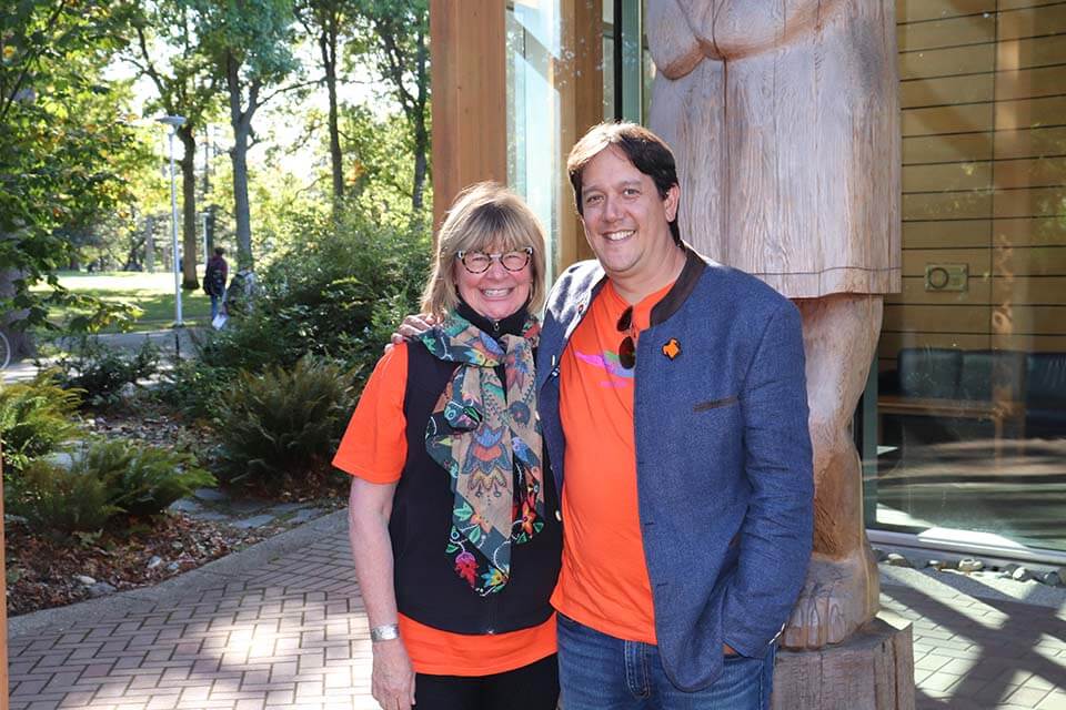 UVic visual artist Carey Newman (Kwakwaka’wakw/Coast Salish) and UVic Chancellor Shelagh Rogers pose in their orange shirts in front of the First Peoples' House on the UVic campus.