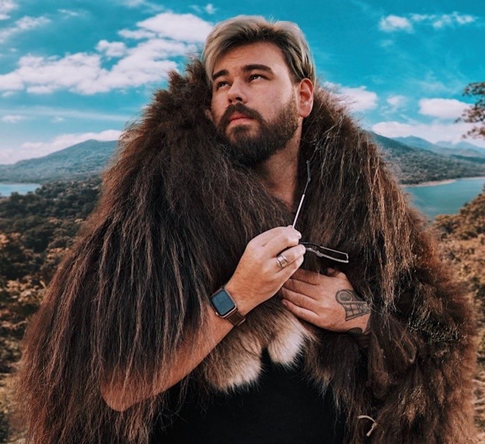 Young Indigenous man with moustache and beard and blond hair pictured standing outdoors with mountains and ocean in the background. He is holding a pair of sunglasses in his hand and looking off camera. A partial tattoo is visible on his left hand and he is wearing a dark brown fur draped over his shoulders and a black t-shirt.