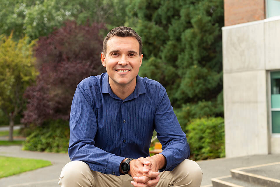 Alan McConnachie sits in front of building at UVic, smiling.