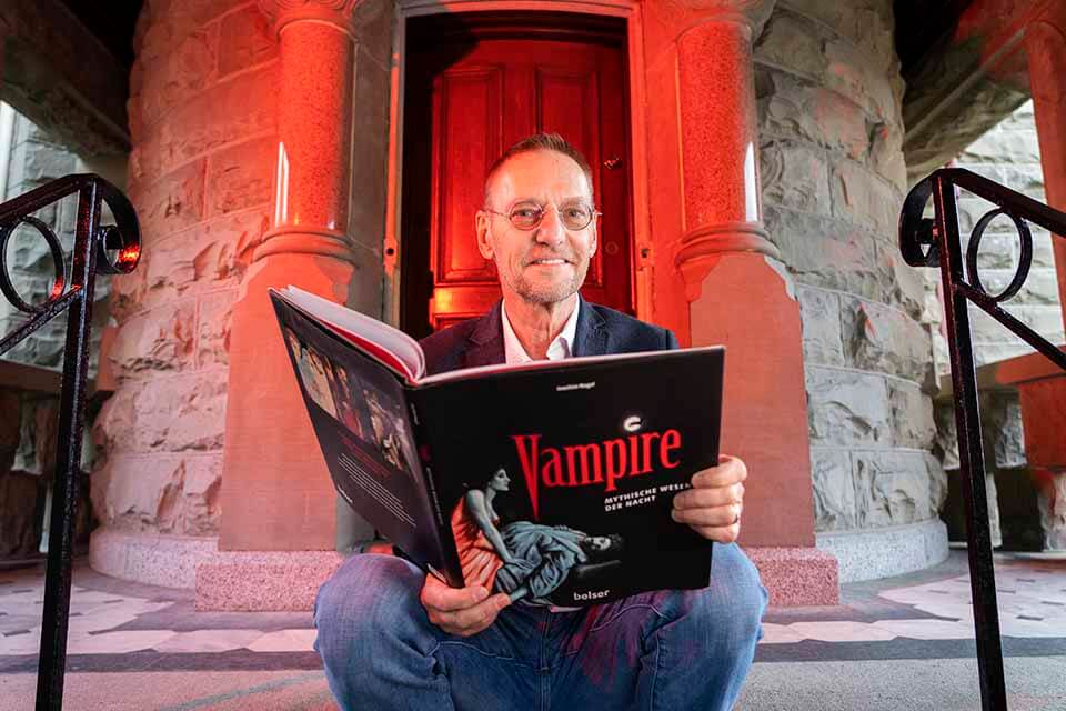 Peter Golz sits outside a medieval-looking stone building, the doorway lit up in red. He holds a book with a cover that reads 