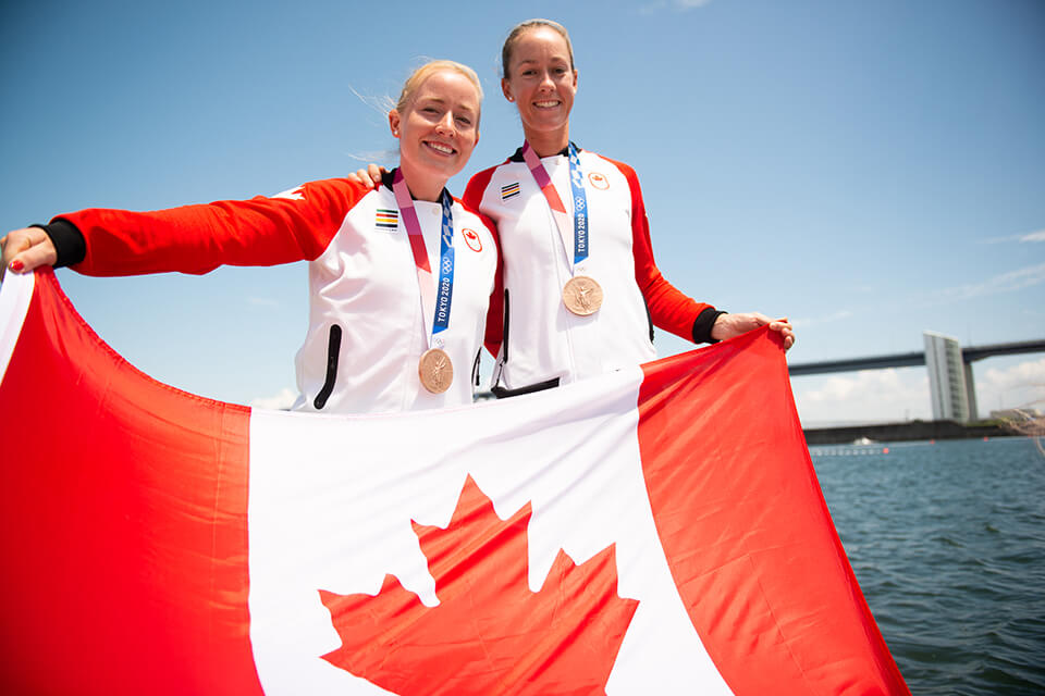 UVic rowing alumna Caileigh Filmer poses with her rowing partner Hillary Jansens. They wear their newly awarded Olympic bronze medals and hold a large Canadian flag. 