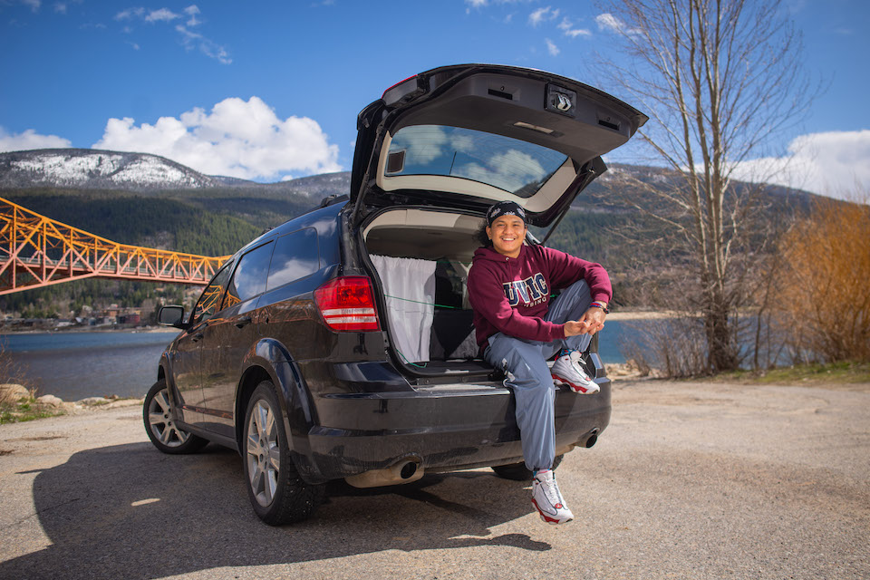 James smiles while leaning on back open trunk of a black SUV that has curtains pulled along the back inside for privacy.