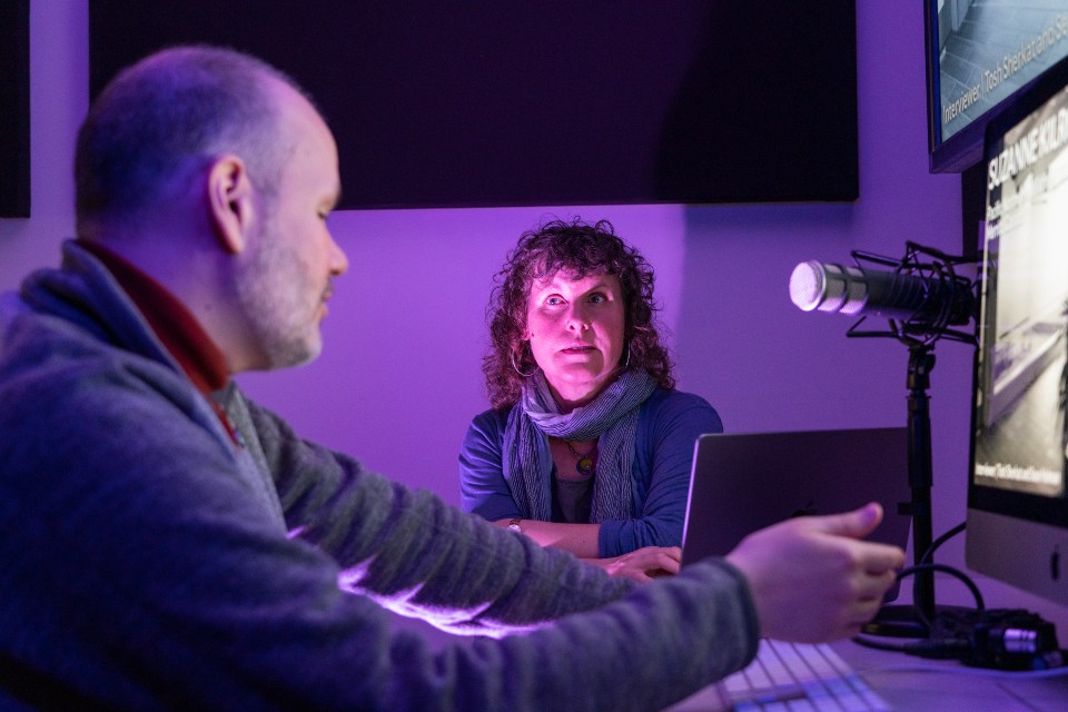 A person facing the camera is listening to a person partly out of the frame. They sit at a desk where there are two monitors and a microphone.