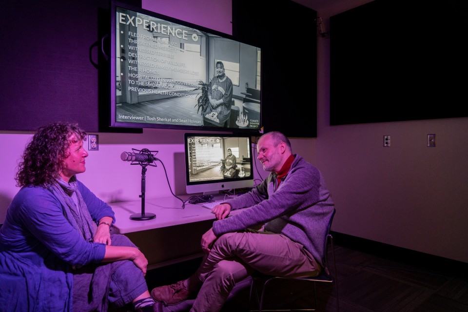 A person facing the camera is listening to a person partly out of the frame. They sit at a desk where there are two monitors and a microphone.