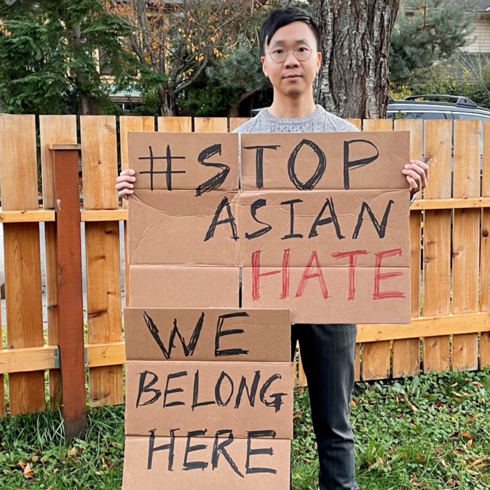 UVic researcher Nigel Lou holds an anti-Asian racism sign, with leafy neighbourhood backdrop