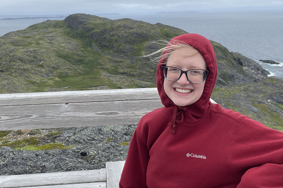 Sophie Pavlik, wearing a red hoodie, smiles at the camera with grassy hill and ocean behind her.