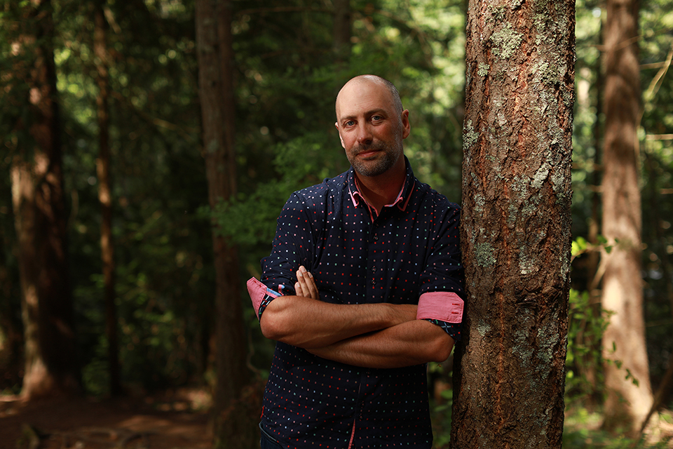 Man in collared shirt leaning up against tree in a forest.