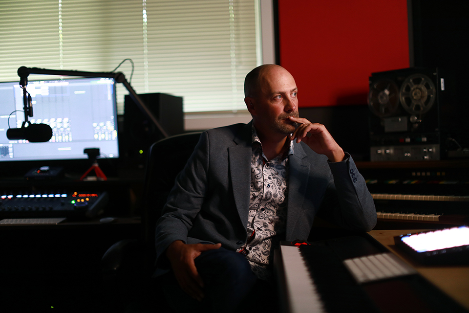 Man sitting in a darkened sound studio beside a microphone, looking off to the right of the frame